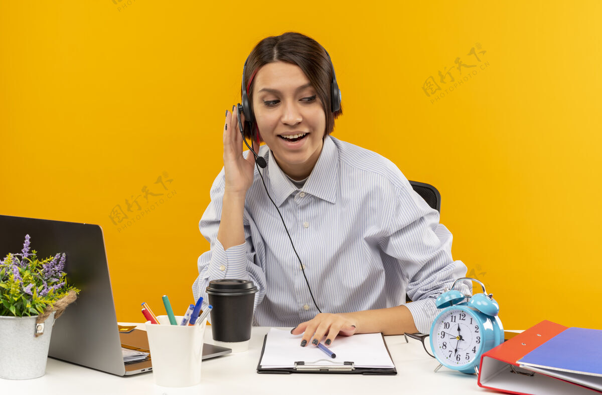 电话印象深刻的年轻呼叫中心女孩戴着耳机坐在办公桌旁 把手放在耳机上 看着一边孤立的橙色印象年轻女孩