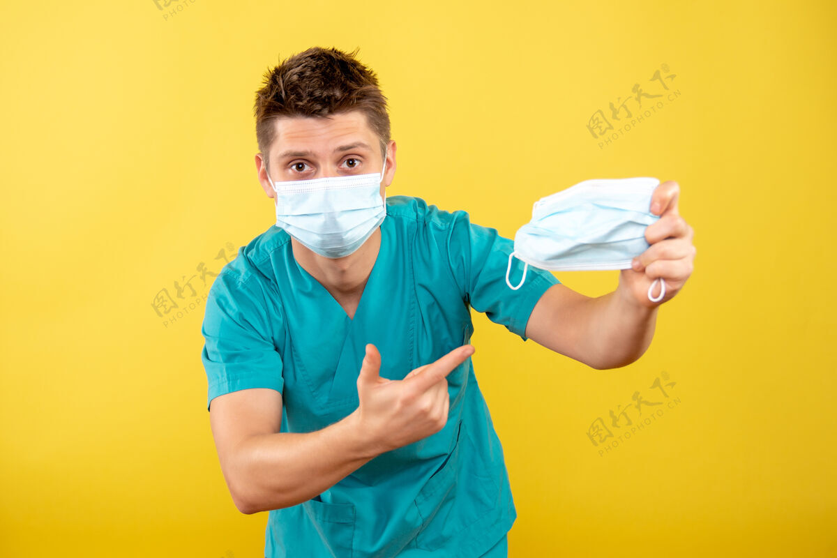 医学正面图年轻男医生穿着医疗服 戴着无菌口罩 拿着黄色背景上的其他口罩人持有套装