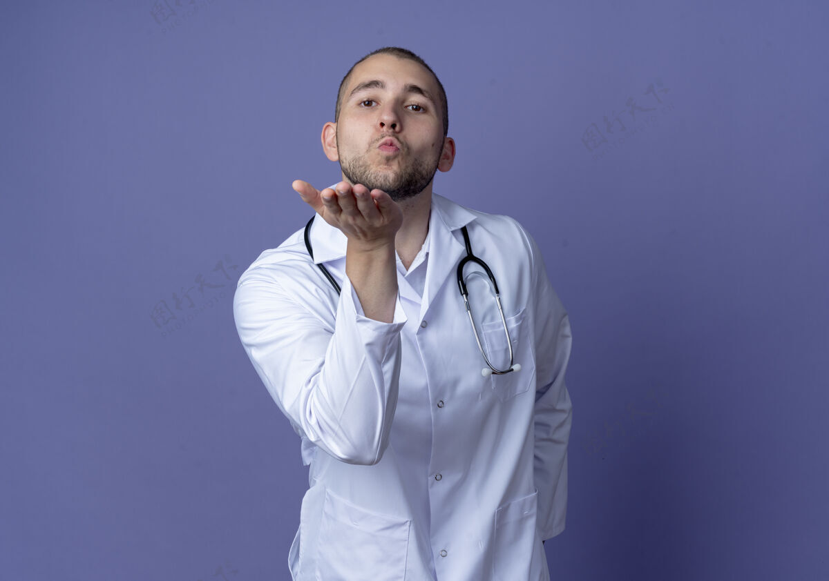 听诊器自信的年轻男医生穿着医用长袍和听诊器送吹吻 看着紫色的复制空间孤立发送长袍吹