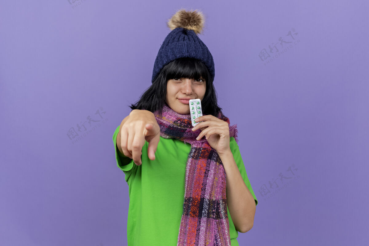 年轻戴着冬日帽 戴着围巾的年轻病态白人女孩高兴地抚摸着嘴唇 手里拿着一包胶囊 看着并指着隔离在紫色背景上的相机 还有复印空间指向请相机