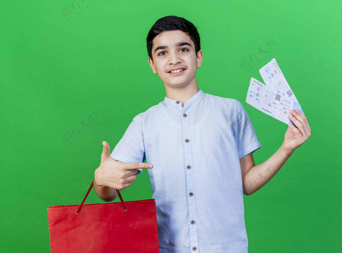 男孩高兴的年轻高加索男孩拿着购物袋和机票指着票看着隔离在绿色背景下的相机年轻人票指向