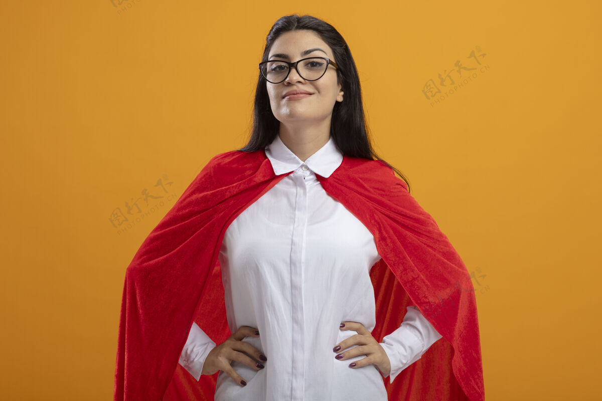 超级英雄高兴的年轻白种人超级英雄女孩戴着眼镜 手放在腰上 看着隔离在橙色背景下的相机女孩腰相机
