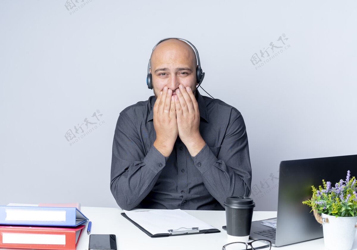 嘴自信的年轻秃头呼叫中心男子戴着耳机坐在办公桌旁 手放在嘴上 与白色隔离工具工作手