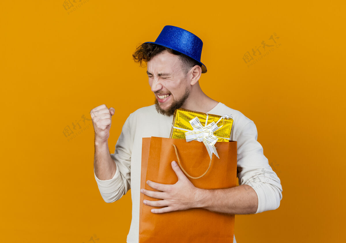 帅气快乐的年轻英俊的斯拉夫党家伙戴着党的帽子拿着礼品盒在纸袋做是的手势孤立在橙色背景与复制空间抱着盒子手势