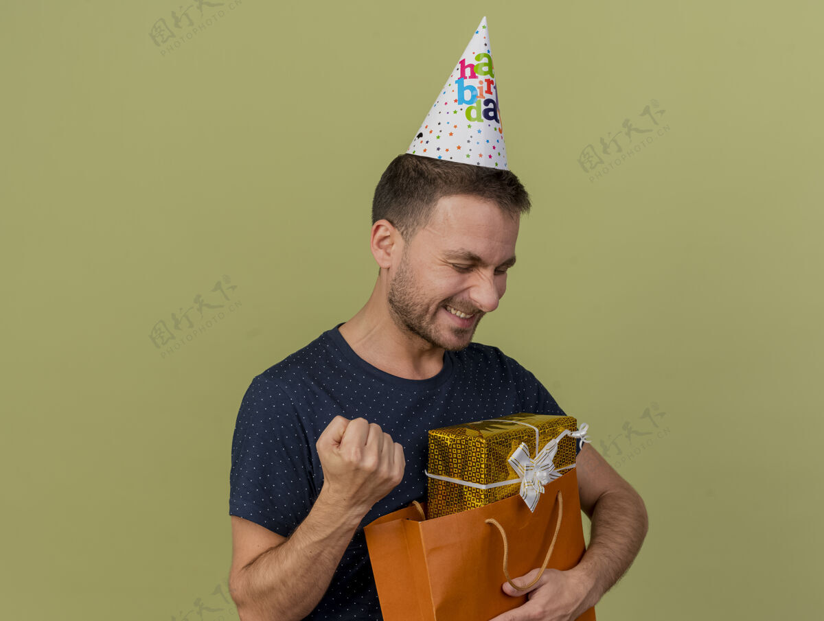 持有戴着生日帽的快乐帅哥高加索人握紧拳头 将礼品盒放在橄榄绿背景上的纸购物袋中 并留有复印空间保持包礼品