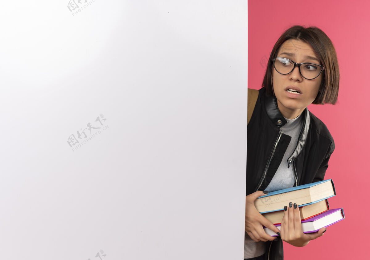 背焦急的年轻女学生戴着眼镜 背着书包 从后面望着侧面 白色的墙上隔离着粉红色的复印空间抱包空间