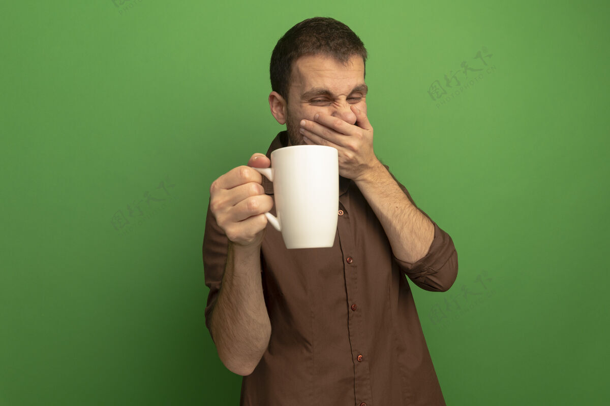 手恼怒的年轻白人男子孤立地站在绿色的怀抱里 看着一杯茶 手放在嘴边 背景是复印空间嘴杯子恼怒