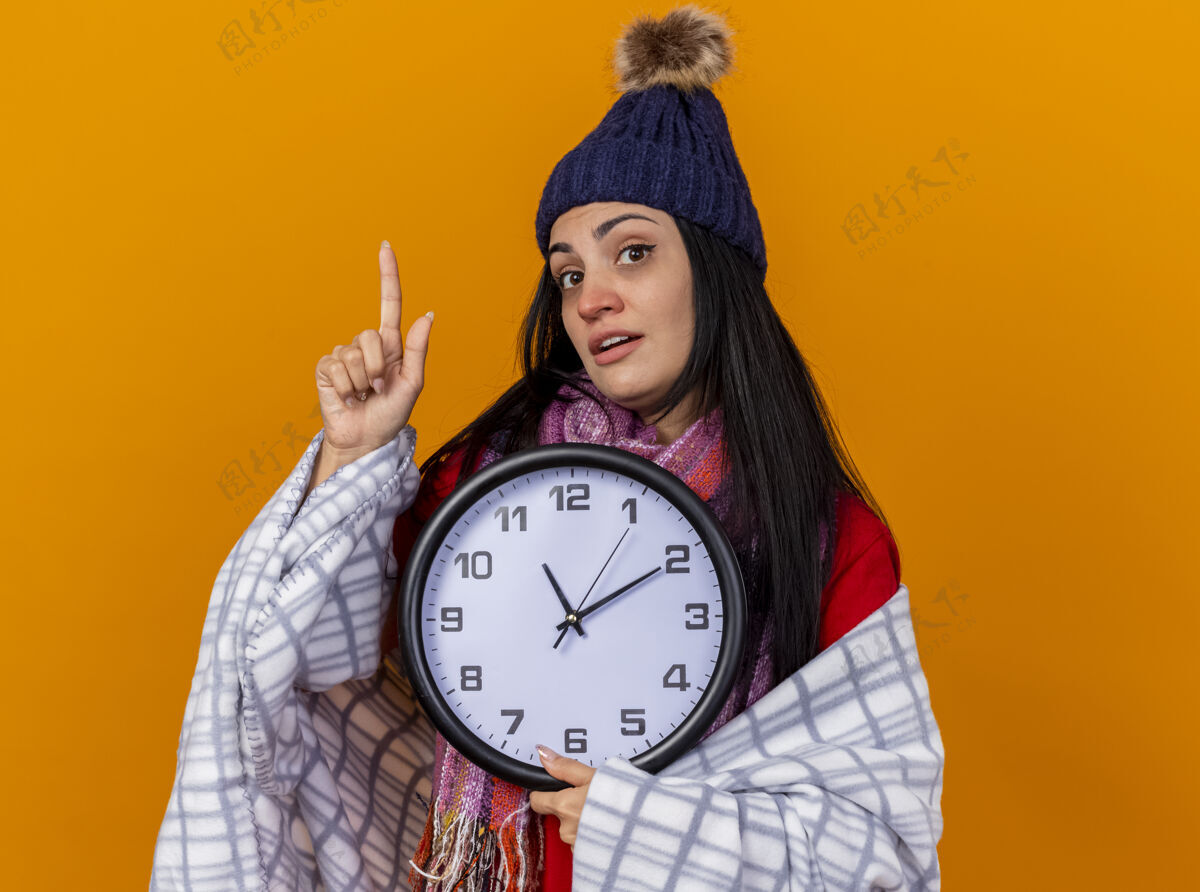 格子布印象深刻的年轻白种人生病的女孩戴着冬天的帽子和围巾包裹在格子布拿着时钟看着相机指向橙色背景上孤立的复制空间指向印象相机