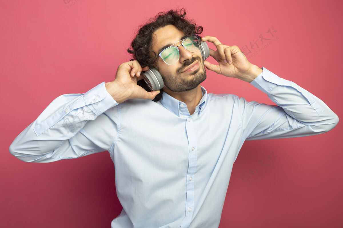眼睛高兴的年轻英俊的白人男子戴着眼镜和耳机抓着耳机听音乐闭上眼睛隔离在深红色的背景请耳机音乐