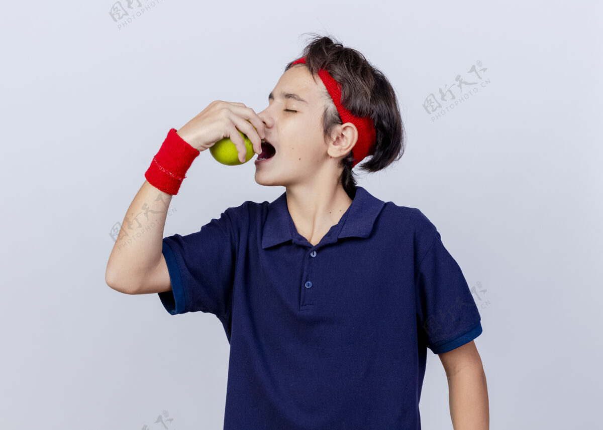 背带年轻英俊的运动男孩戴着头带和护腕 戴着牙套 头对侧地咬着苹果 闭着眼睛 隔离在白色背景上帅气牙齿腕带