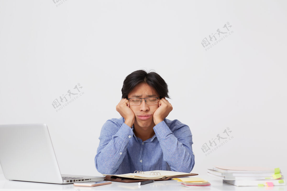 男人疲惫困倦的亚洲年轻商人戴着眼镜 头枕在手上 坐在白墙那边的桌子旁睡觉无聊手桌子