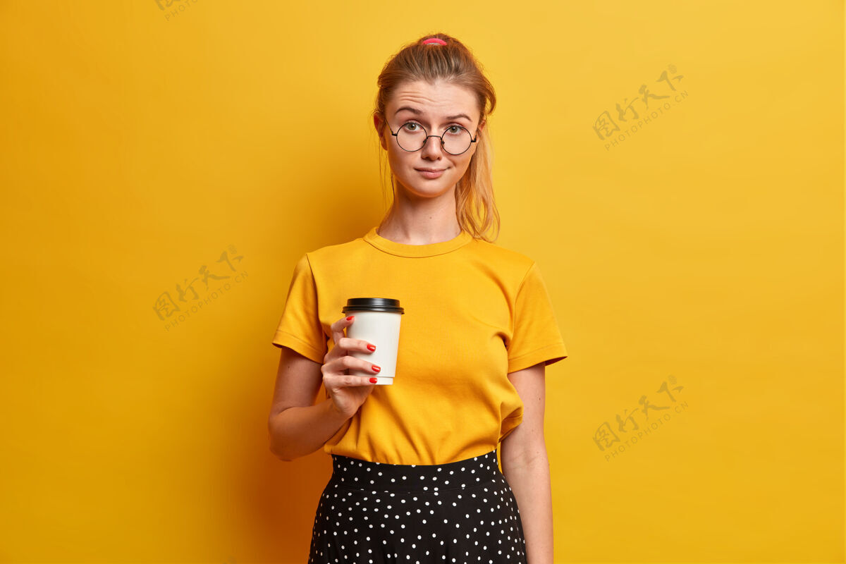 欧洲人严肃漂亮的女孩和新朋友闲暇时拿着外卖咖啡饮料 看起来不开心地穿着黄色t恤眼镜站在室内休闲魅力咖啡因