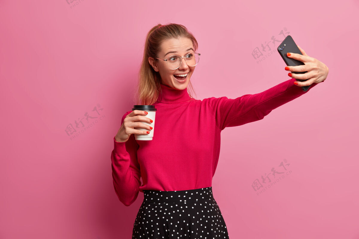 在线乐观的千禧一代女孩录制视频 在现代智能手机上自拍 通过手机应用打电话给朋友 喝咖啡去 用清爽的饮料开始新的一天积极杯子通信