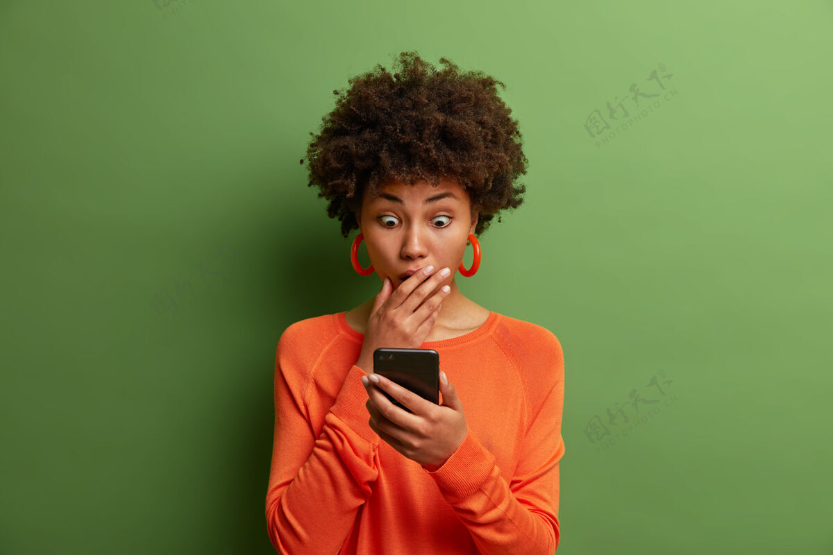 反应惊诧的成年女子大吃一惊 盯着智能手机屏幕 在网站上读着令人震惊的新闻 穿着橙色套头衫 眼睛被窃听 嘴巴被遮住天哪 太可怕了Omg惊人手机