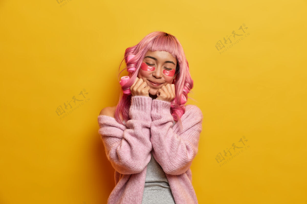 完美满心欢喜的粉红头发的亚洲女人把手放在下巴下 闭上眼睛 涂上发卷贴片纯净满意