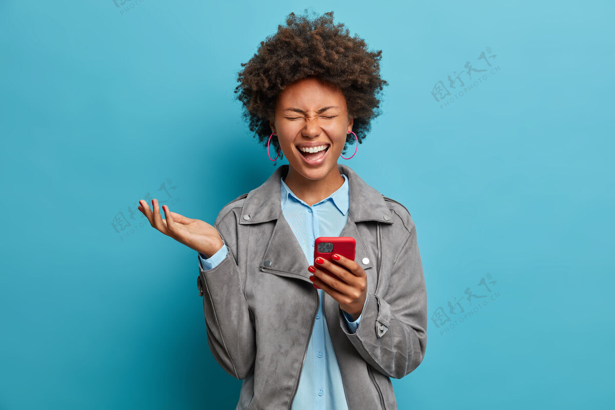 短信喜出望外的卷发民族女子 笑着在智能手机上看搞笑视频 闭上眼睛举手 心情很好 穿着灰色休闲夹克 摆姿势微笑年轻民族