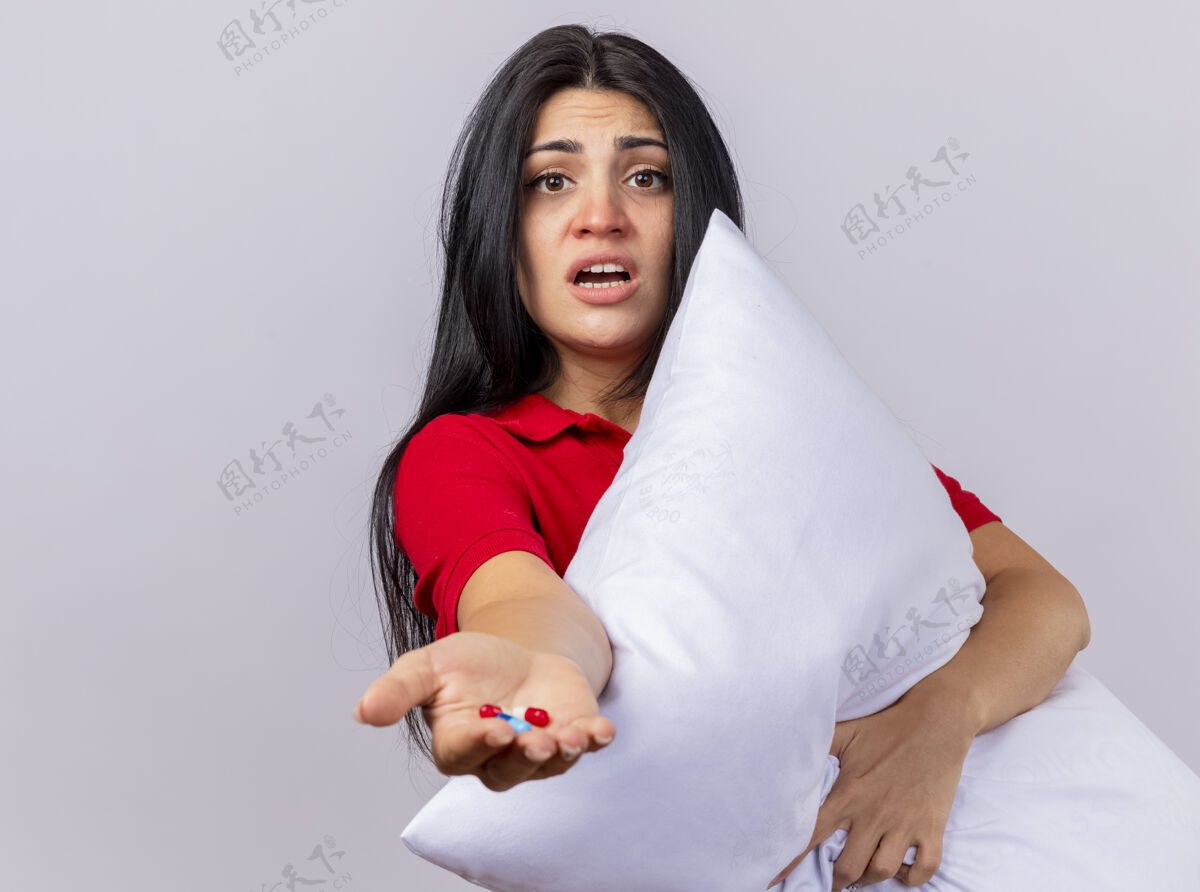 印象印象深刻的年轻白种人生病的女孩抱着枕头看着相机伸展胶囊对相机隔离在白色背景与复制空间年轻拥抱空间