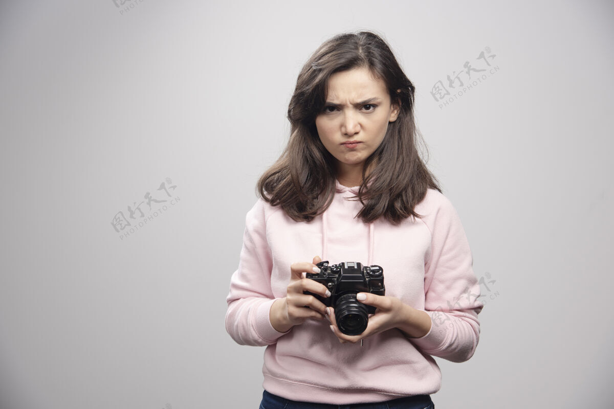 女人愤怒的摄影师拿着相机站在灰色的墙上女孩女性摄影师