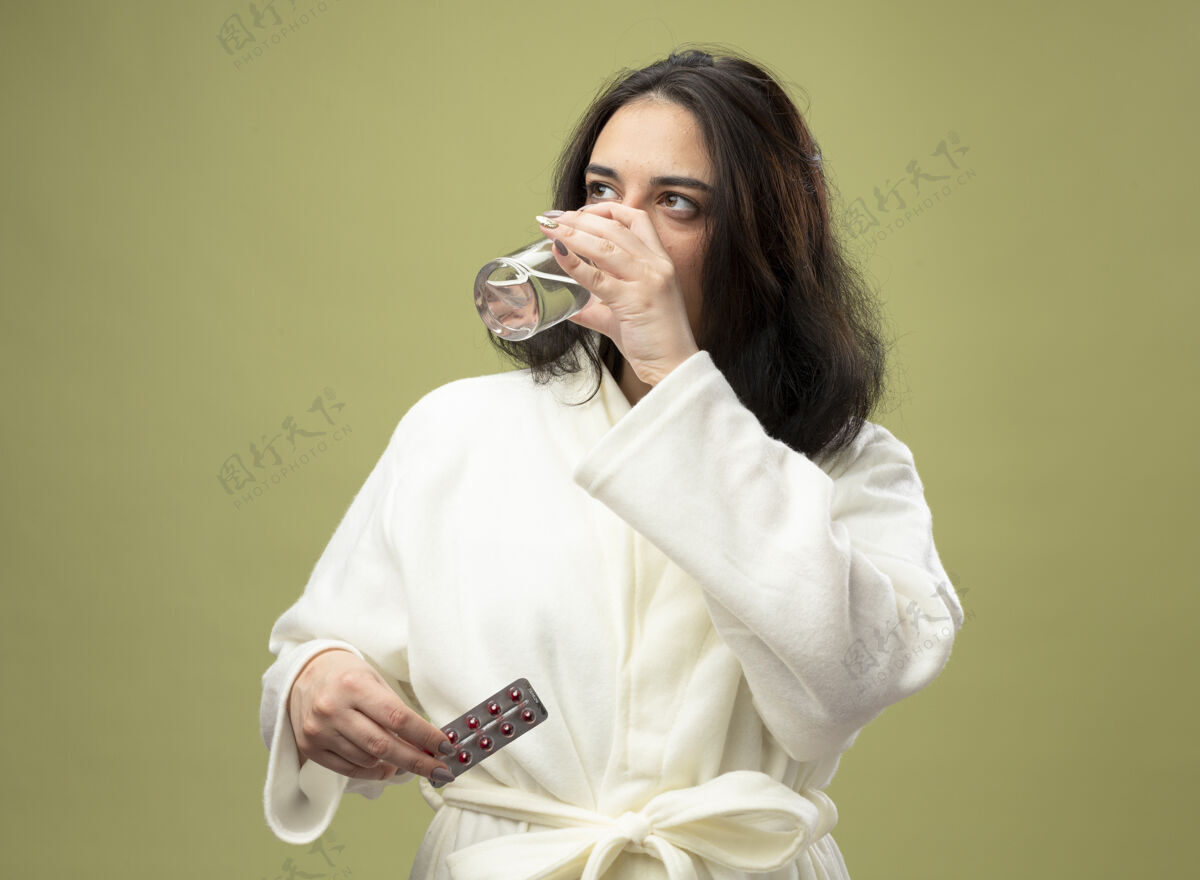 壁板年轻的白种人生病的女孩穿着长袍 喝着一杯水 手里拿着一包药丸 看着一边孤立的橄榄绿背景年轻疾病女孩