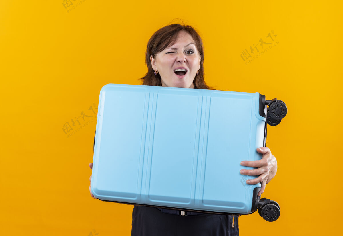 眨眼眨眼的中年旅行家妇女拿着手提箱站在隔离的黄色墙上手提箱女人中年人