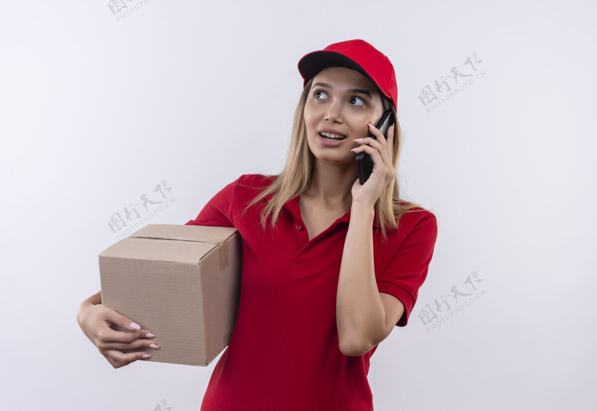 说话看着身边高兴的年轻送货女孩穿着红色制服 戴着帽子拿着盒子 隔着白墙讲着电话壁板穿帽子