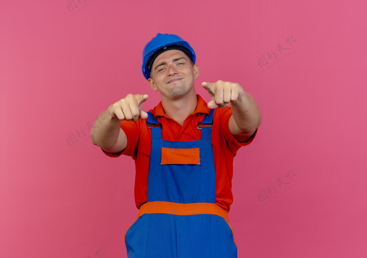 头盔高兴的年轻男子建设者穿着制服 戴着安全帽指着镜头上的粉红色请年轻人男