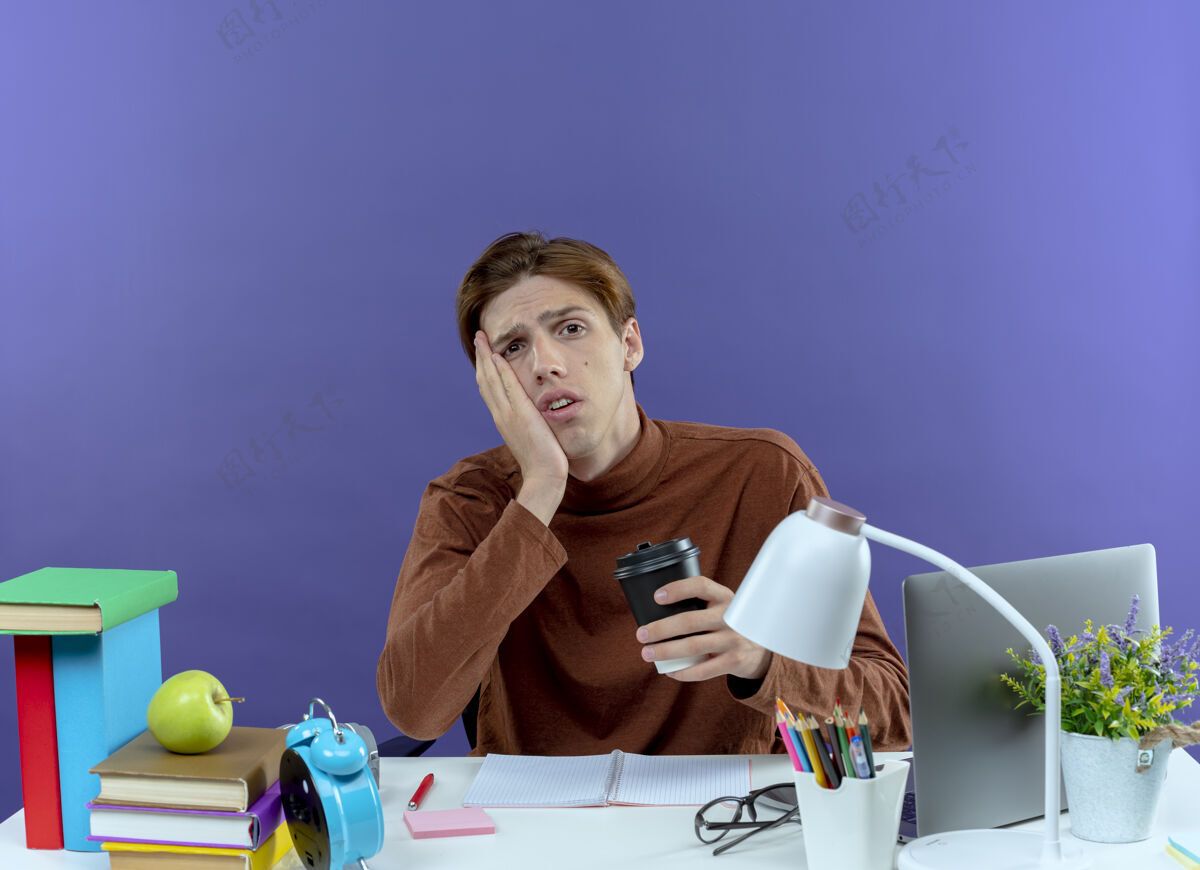 坐着悲伤的学生男孩坐在书桌旁 手里拿着学习用具 手里拿着一杯咖啡 手放在紫色的脸颊上紫色抱着年轻
