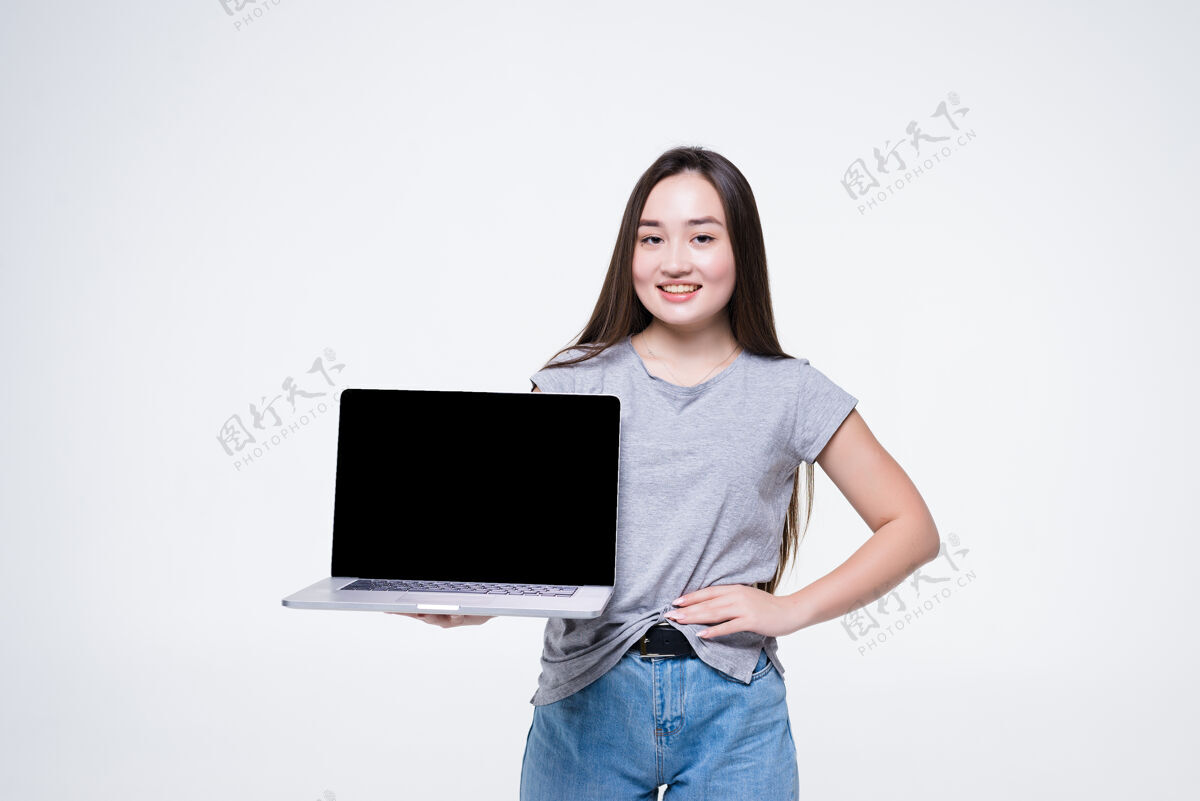 持有一个快乐的年轻亚洲妇女的肖像指着空白屏幕笔记本电脑 而坐在白墙隔离积极肖像人