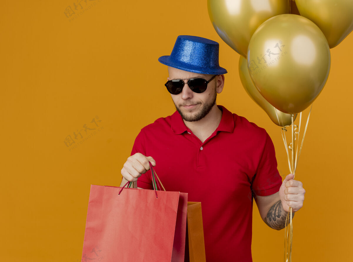 人愁眉不展的年轻帅哥戴着墨镜和派对帽拿着气球和纸袋孤立在橙色的墙上太阳镜气球包