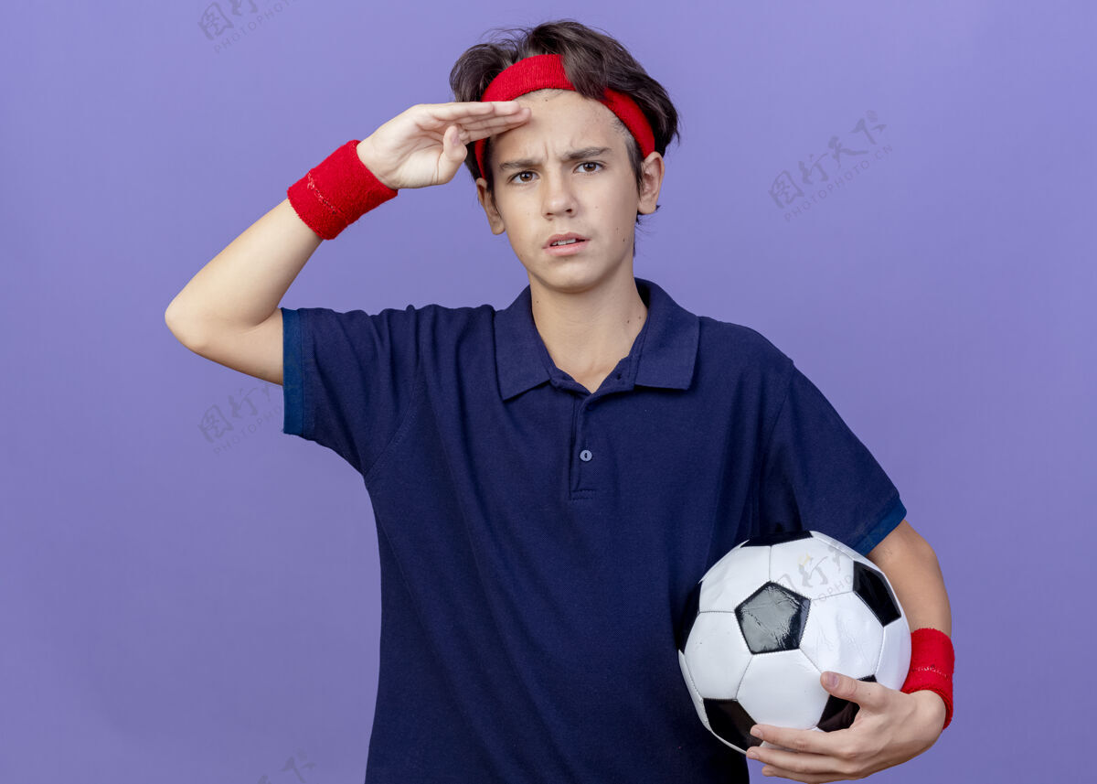 人皱眉的年轻英俊的运动男孩戴着头带和戴着牙套的腕带拿着足球看着前面 手放在额头上 隔离在紫色的墙上帅气足球感觉