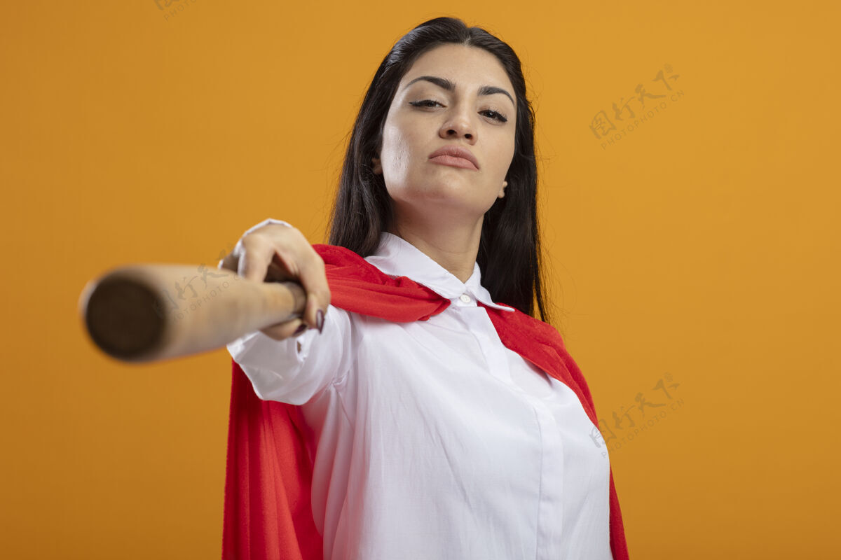 超级英雄自信的年轻女超人向前伸出棒球棒 看着橙色墙壁上孤立的前方姿势女人棒球