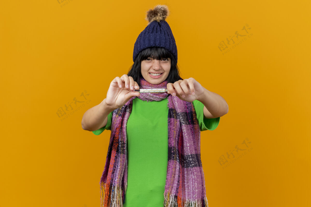 感觉恼怒的年轻生病的女人戴着冬天的帽子和围巾 向前面伸出温度计 看着隔离在橙色墙上的前面人年轻人烦恼