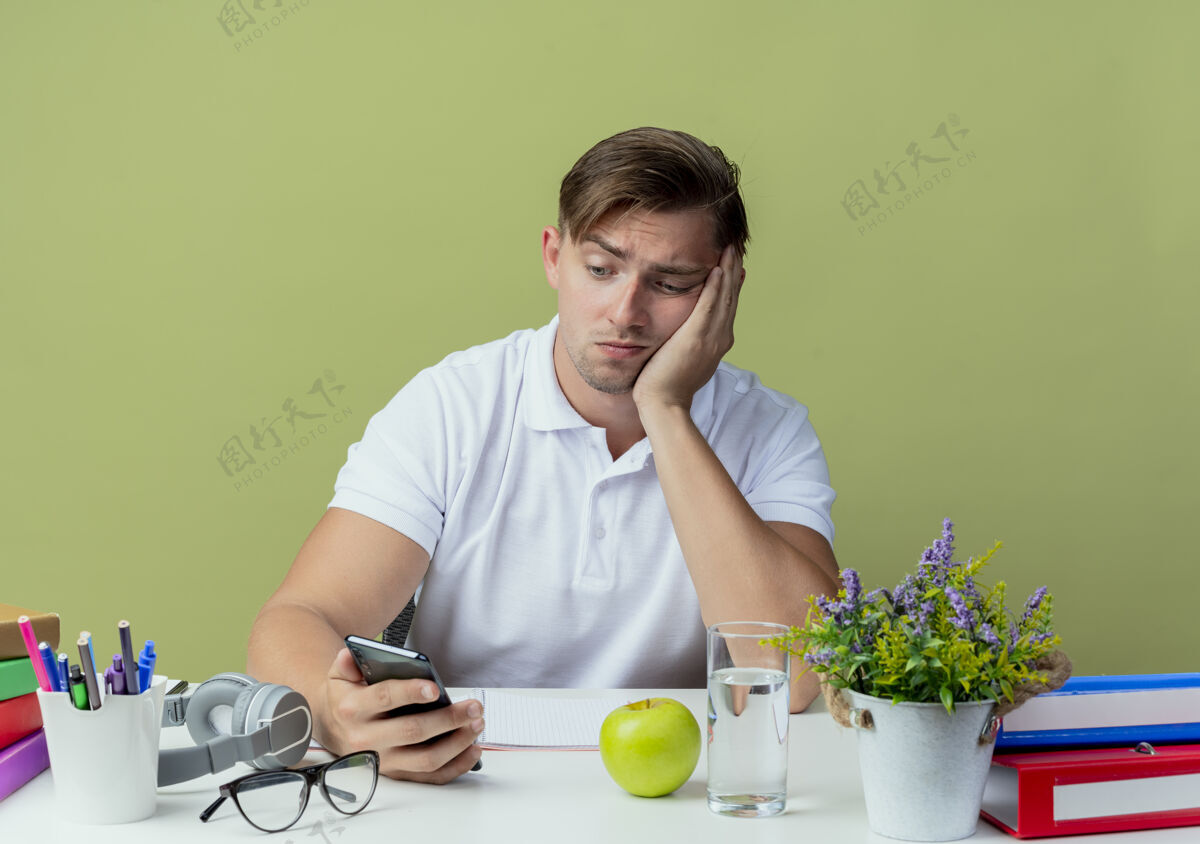 工具悲伤的年轻英俊的男学生坐在书桌旁 手里拿着学习工具 看着橄榄绿上孤立的电话橄榄电话抱着