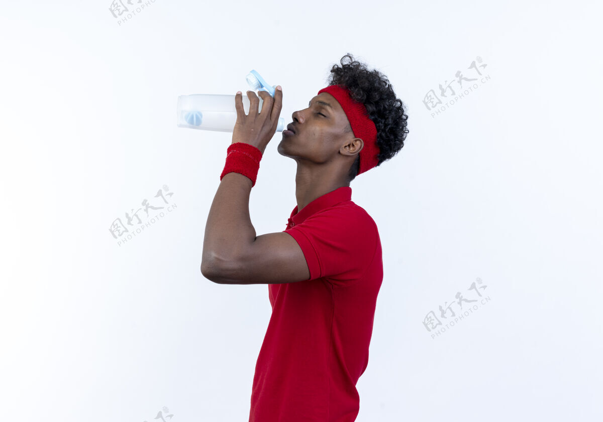 白色站在侧视图年轻的黑人运动男子戴着头带和腕带饮用水从瓶子隔离在白色运动年轻轮廓