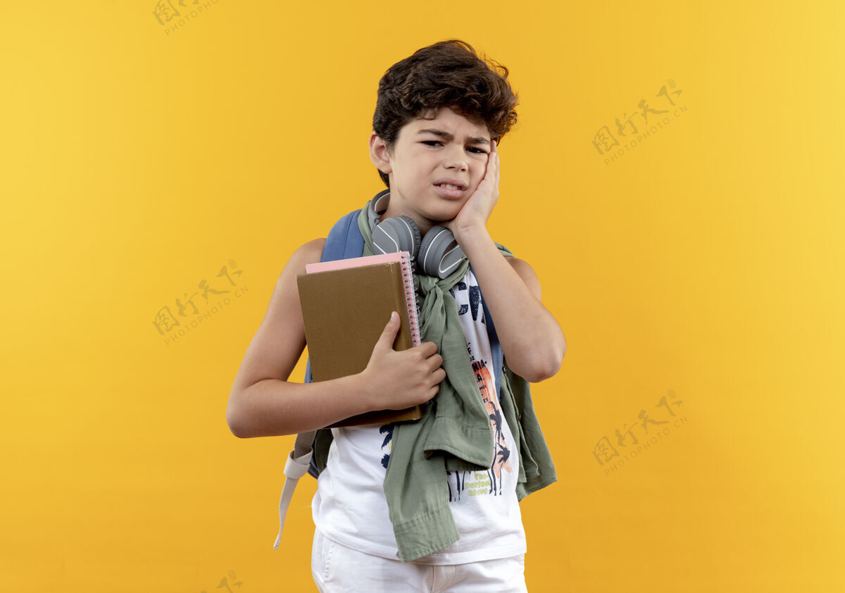 悲伤可怜的小男孩戴着书包和耳机 手里拿着书 手放在脸上 脸上泛着黄色背脸颊表情