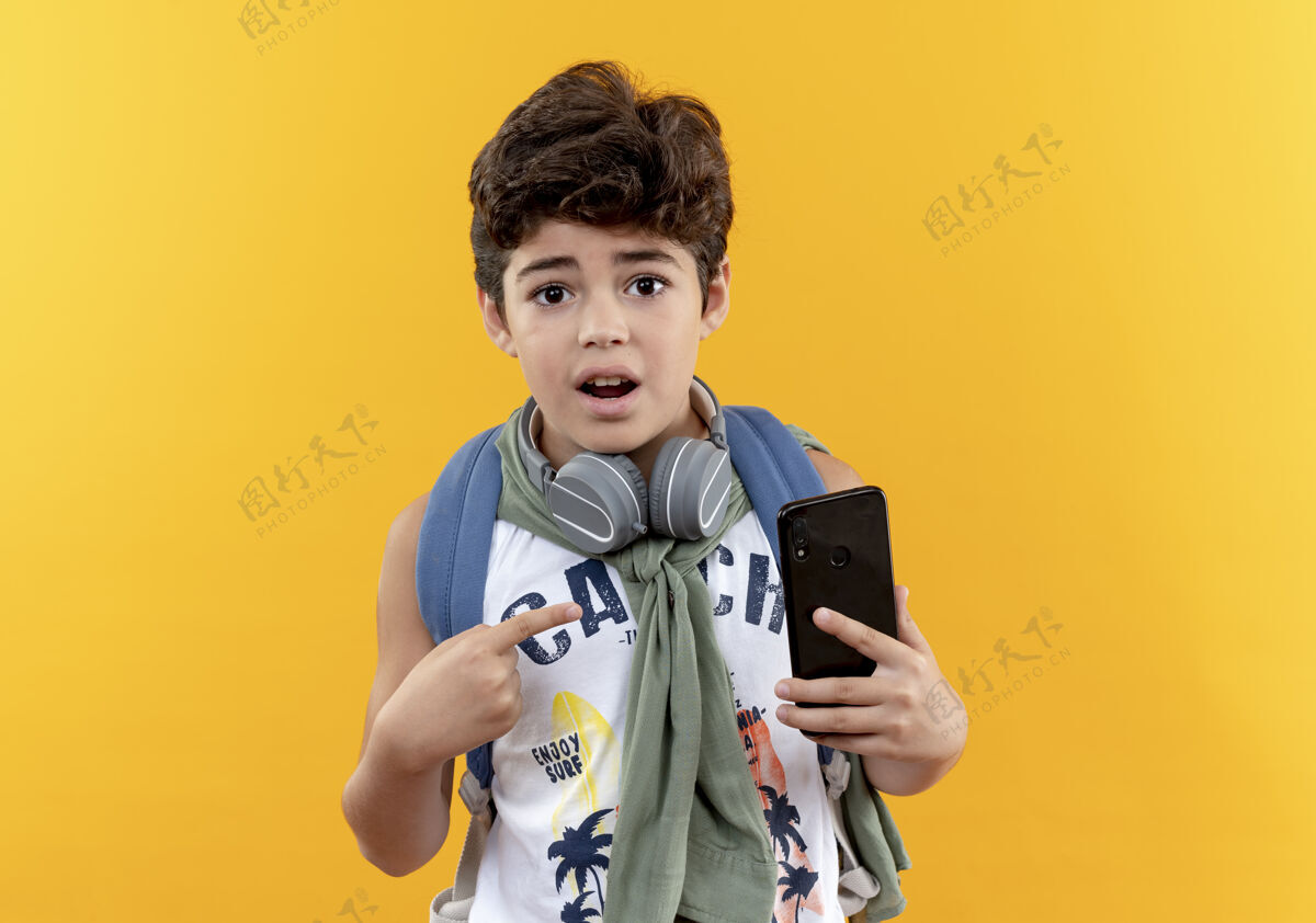 点惊讶的小男生背着书包 拿着耳机 指着隔离在黄色上的电话背表情黄色