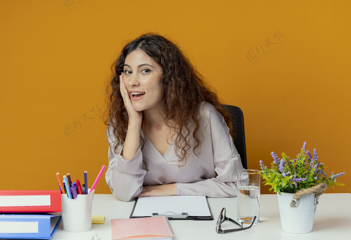 脸颊微笑着年轻漂亮的女上班族坐在办公桌前 手放在橘子色的脸颊上坐着手工人