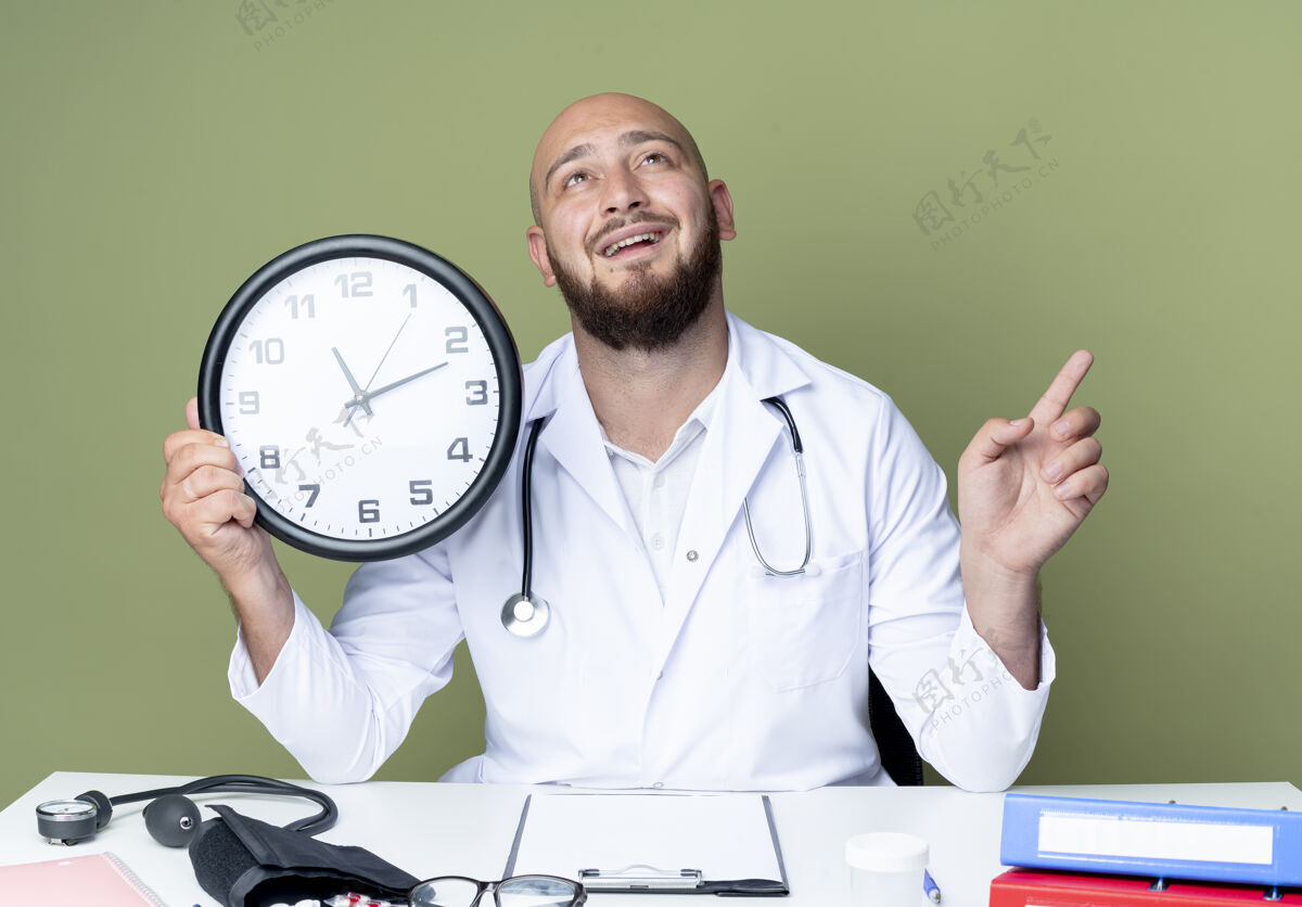 壁板面带微笑的年轻秃头男医生穿着医用长袍和听诊器坐在办公桌前工作长袍秃顶坐着