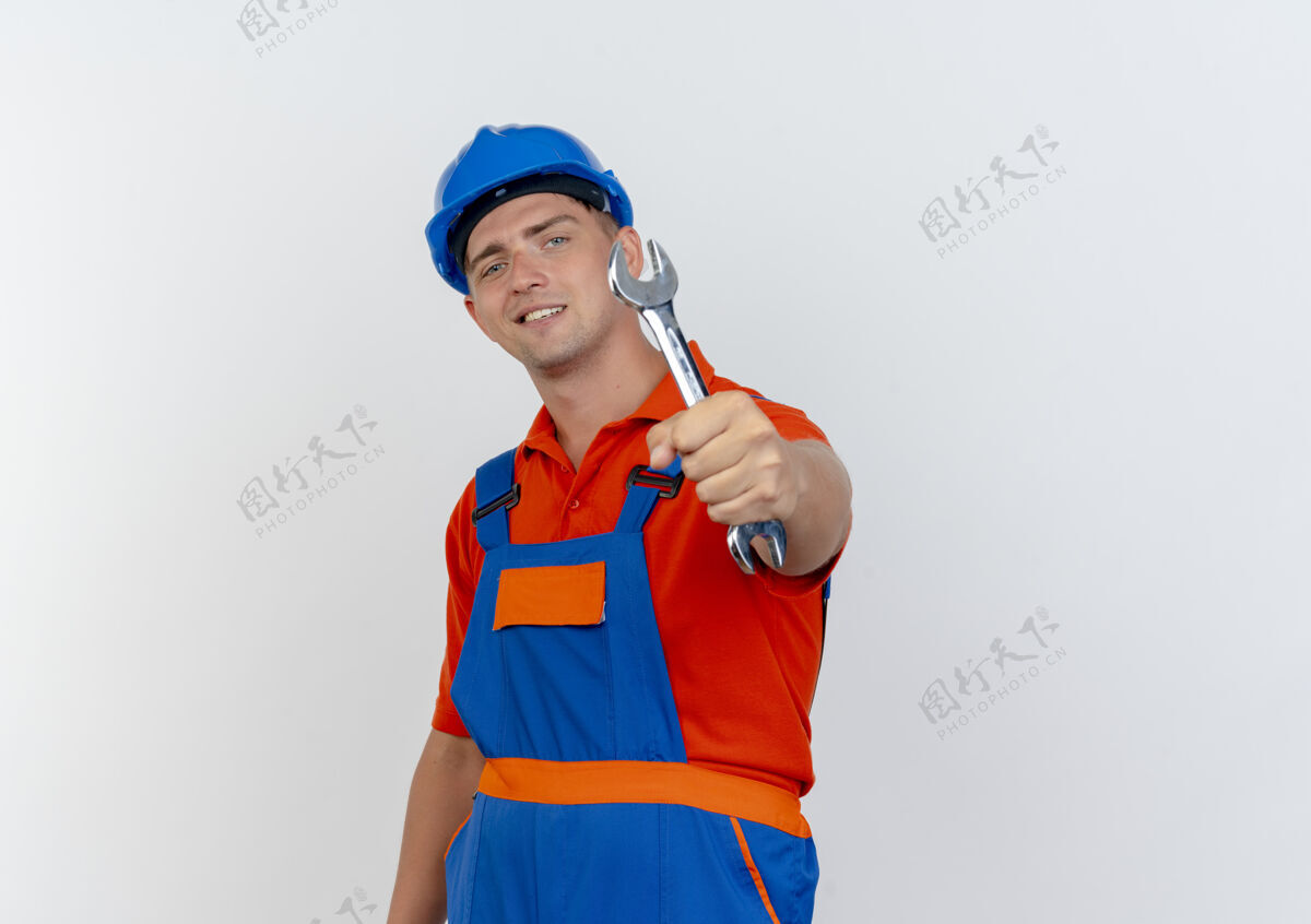 头盔高兴的是年轻的男建筑工人穿着制服 戴着安全帽 手持扳手对着镜头上的白色安全建筑工人制服