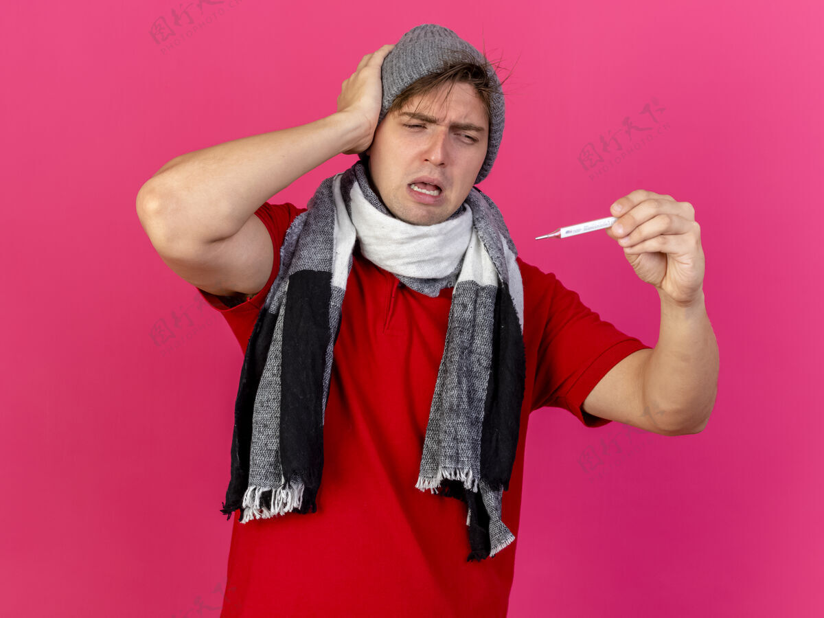 触摸皱着眉头的年轻英俊的金发病患戴着冬天的帽子和围巾拿着温度计看着隔离在粉红色墙上的头市民体温计英俊