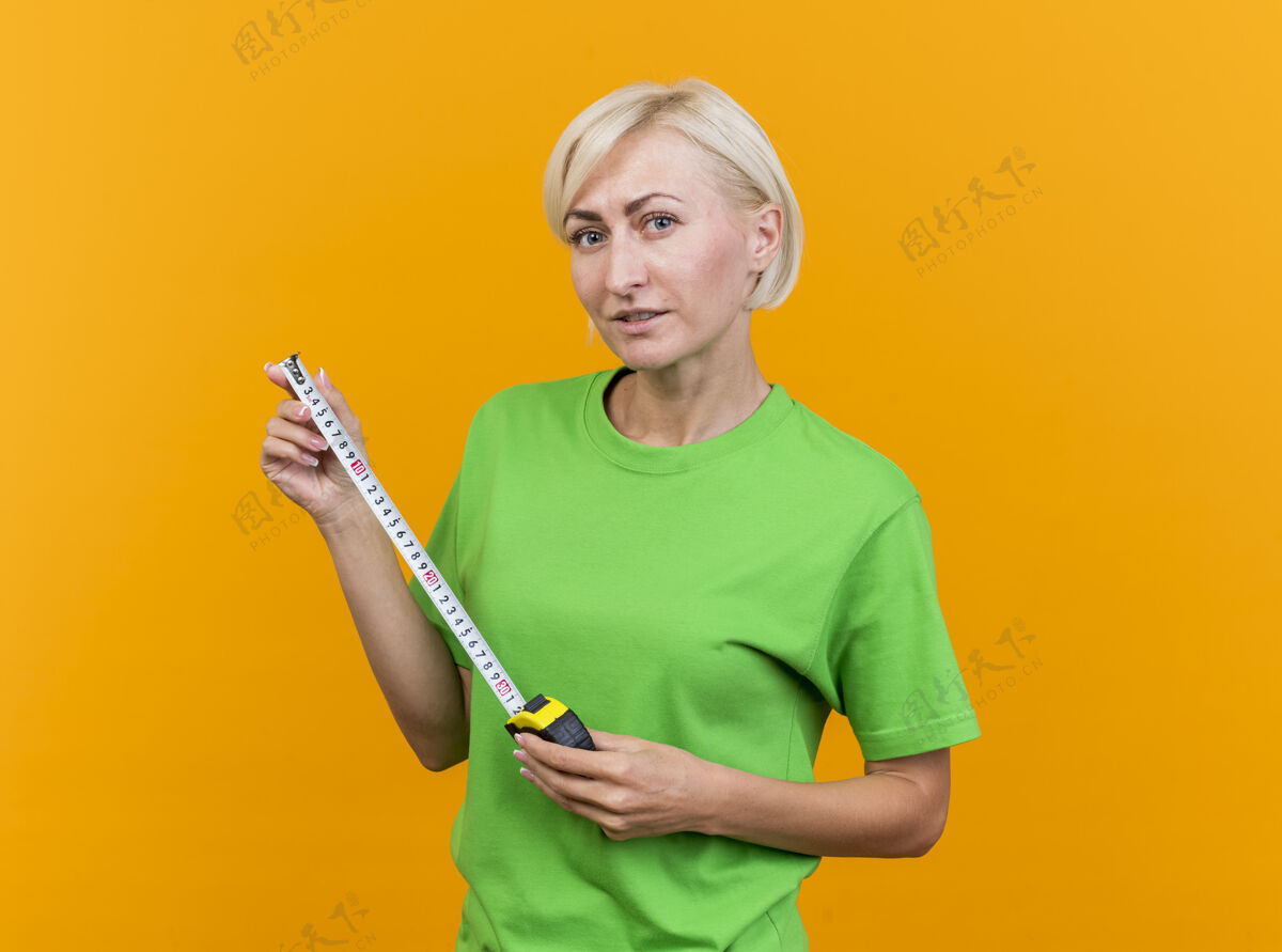 带子自信的中年金发女人看着前面拿着隔离在黄色墙上的磁带表金发站着仪表