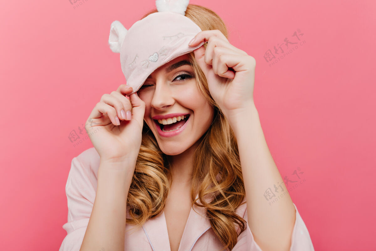 幸福欣喜若狂的金发女士在清晨玩得很开心迷人的欧洲女孩戴着滑稽的眼罩在粉红色的墙上大笑睡衣表情欢乐