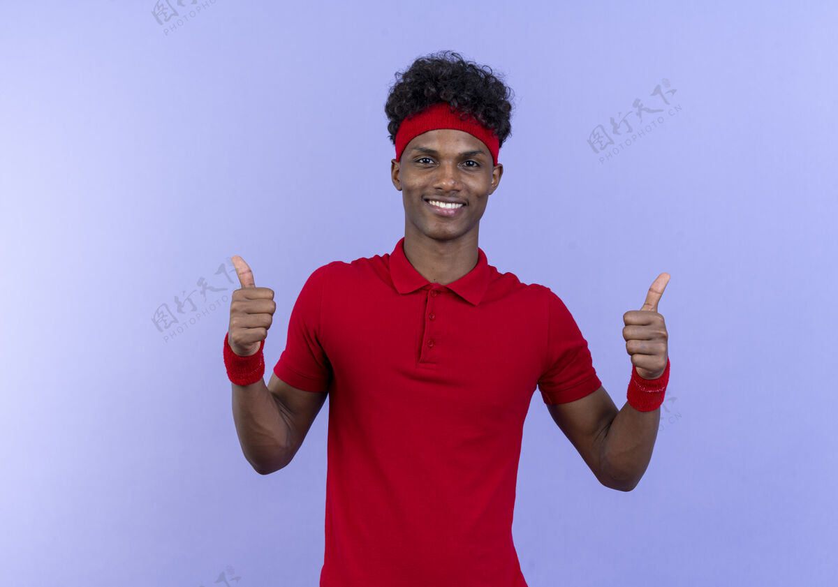 蓝色带着头带和腕带的年轻的美国黑人运动男子微笑着 他的大拇指孤立地竖立在蓝色的地板上男人手势运动