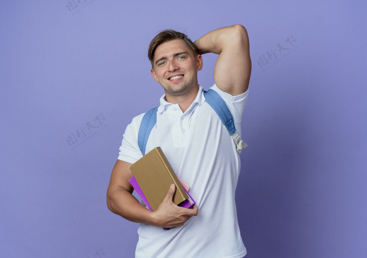 学生面带微笑的年轻帅气男学生背着书包拿着书 手放在脑后 孤零零地躺在蓝色的地板上抱蓝色包