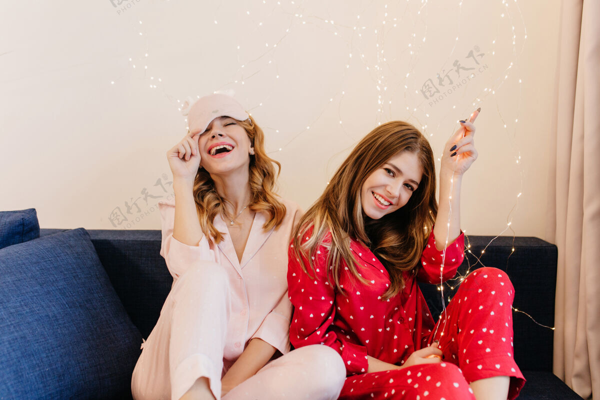 漂亮一头黑发的女孩在沙发上摆姿势 表情很有趣快乐的卷发女士穿着粉色睡衣 戴着睡眠面罩 在蓝色沙发上微笑女士年轻睡衣