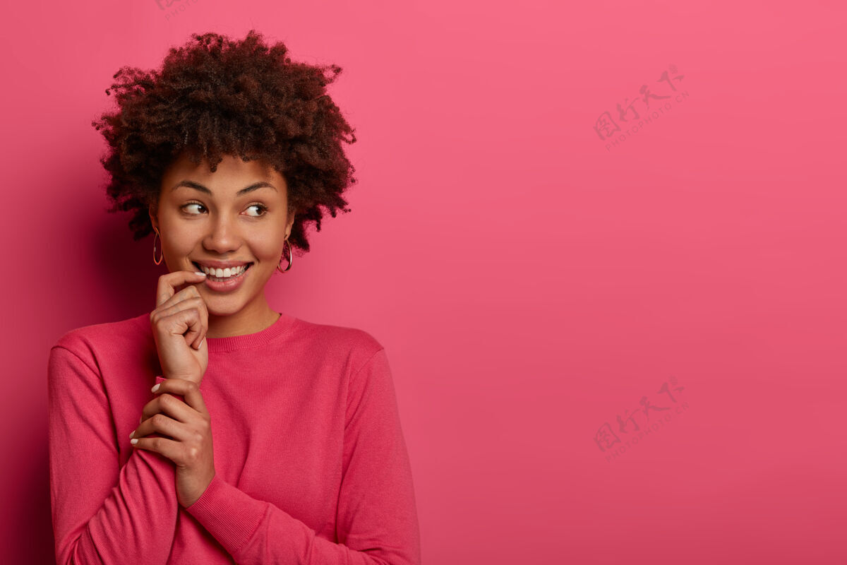 卷发照片中的非洲裔美女 食指靠近嘴巴 注意力集中在一边 表情愉快 注意到一些吸引人的东西 隔离在粉红色的墙上 自由空间欢呼呼吁满意