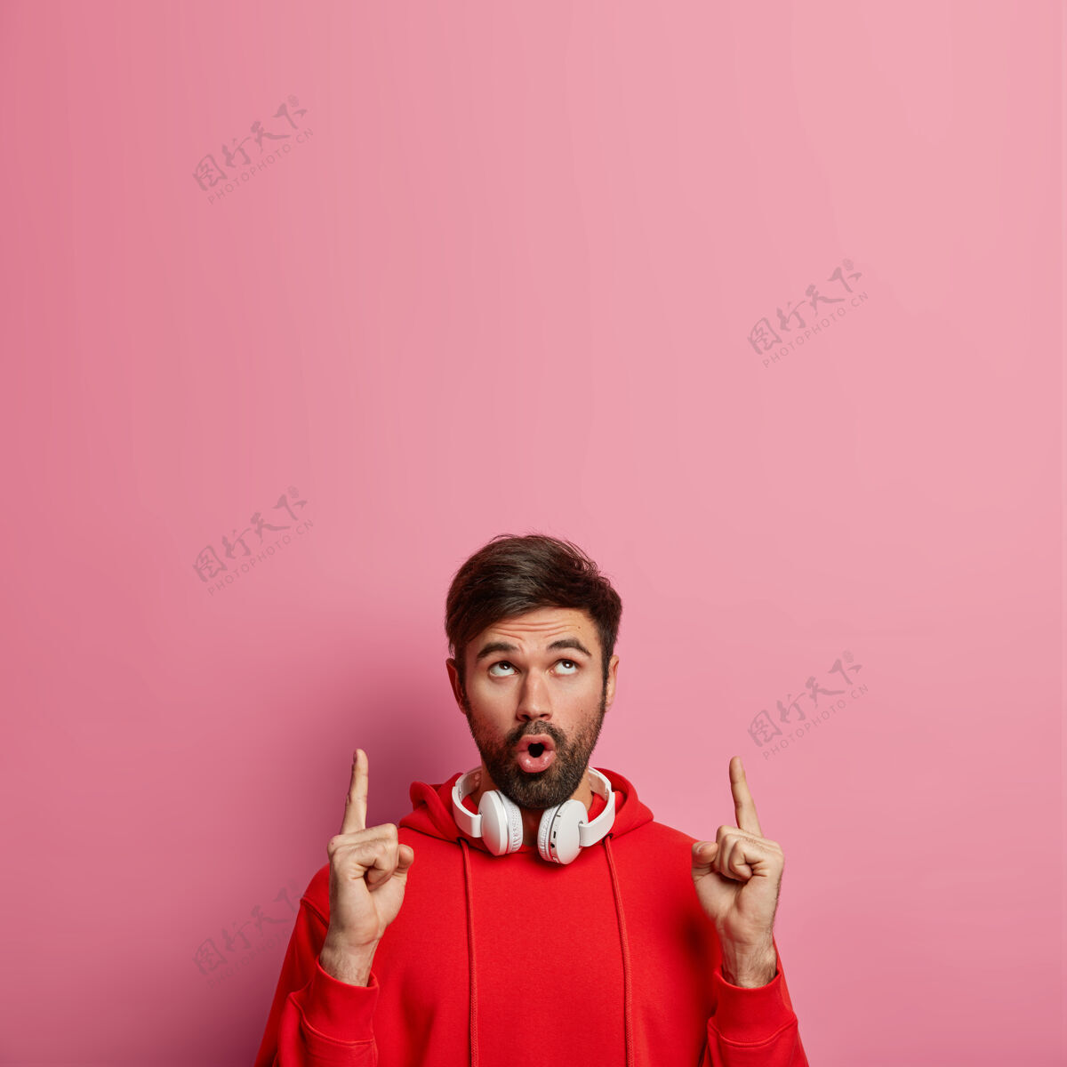 面部迷人印象深刻的胡须男子梅洛曼点以上 显示一些惊人的向上 使用立体声耳机听音乐 喘息的奇迹 隔离在粉红色的墙壁促销概念不刮胡子惊呆男性