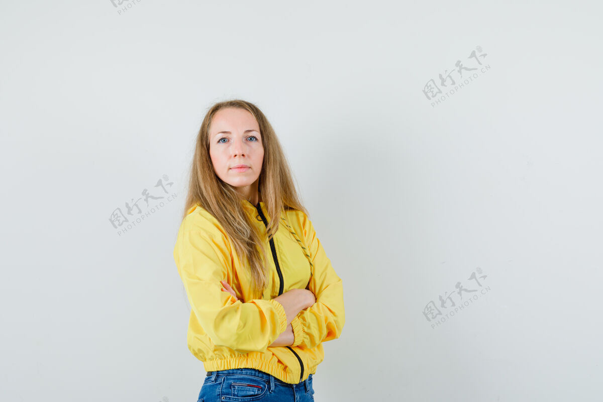 欢呼年轻女子交叉着双臂站着 穿着黄色棒球服和蓝色牛仔裤 冻得发抖 看上去很自信正面图漂亮年轻时尚