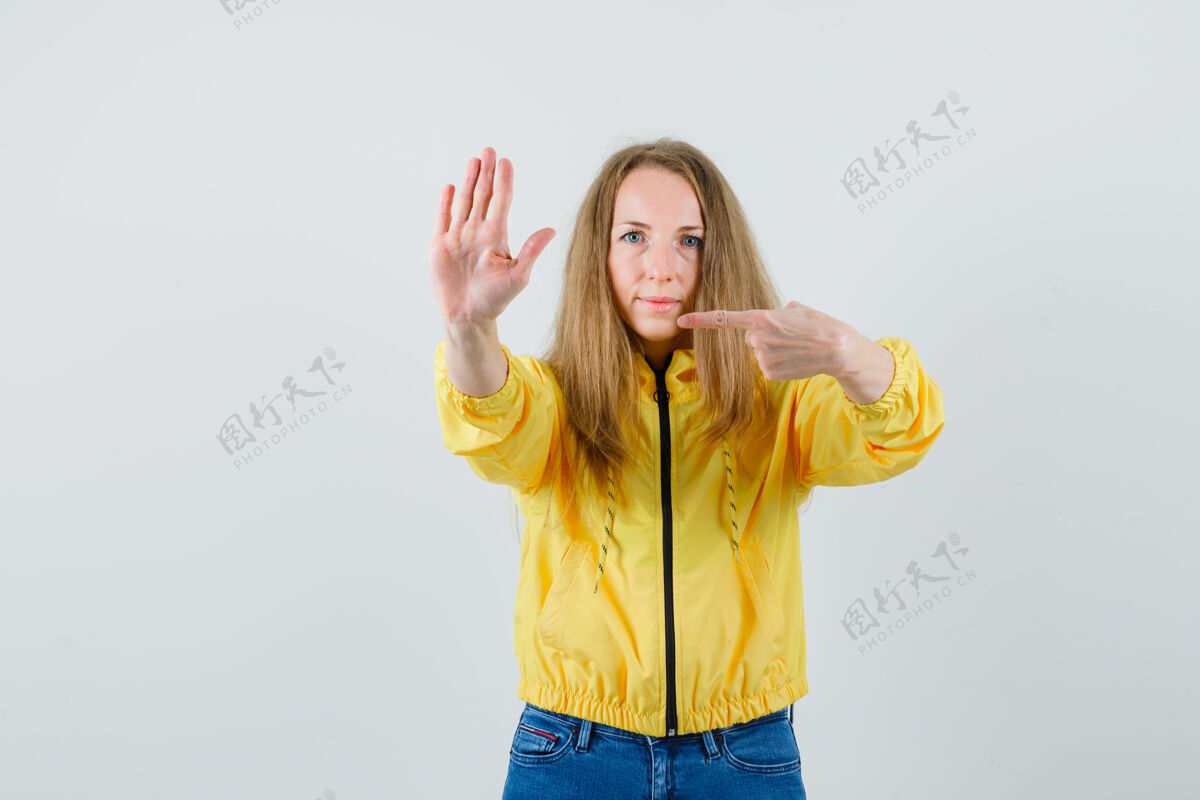 成人身穿黄色棒球服和蓝色牛仔裤的年轻女子 展示着停下来的姿势 指着它 表情严肃 正对着前方欢呼年轻指向