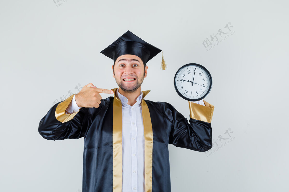 制服年轻人指着挂在墙上的钟 穿着毕业制服 看上去很高兴 正面视图聪明年轻墙壁